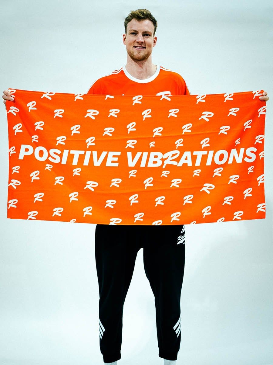 Junger Mann in rotem Sportshirt hält ein orangefarbenes Banner mit weißen Schriftzug "POSITIVE VIBRATIONS" und weißen R-Logos, vor hellem Hintergrund.