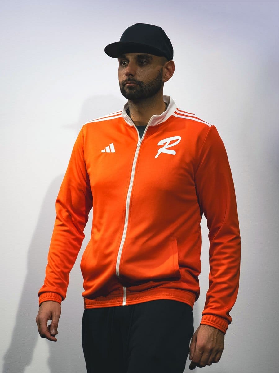 Mann trägt eine orange Sportjacke mit weißen Streifen auf den Schultern, einem weißen Reißverschluss und einem weißen Logo auf der linken Brust. Er trägt eine schwarze Cap.