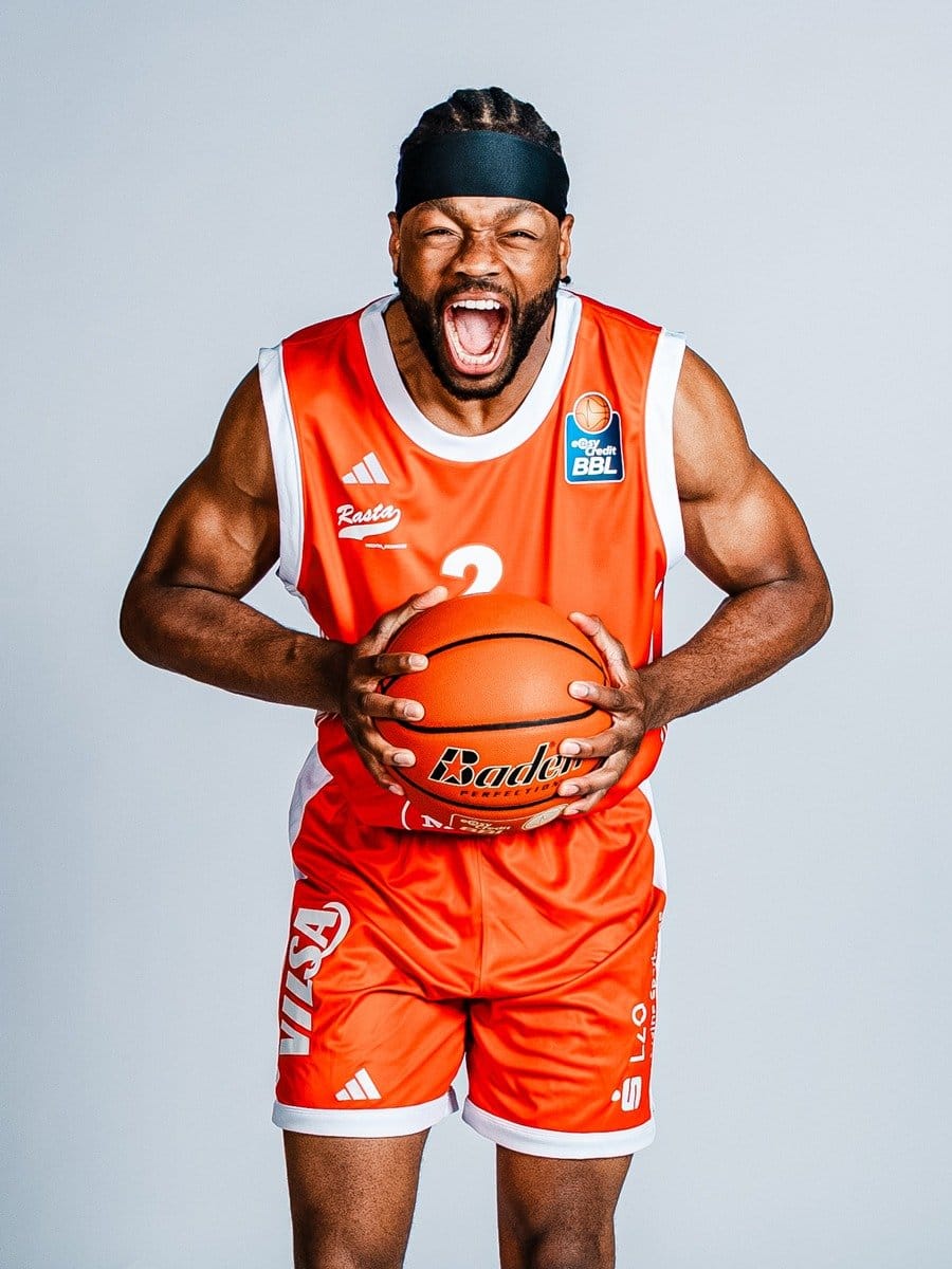 Basketballspieler in rotem Trikot und Shorts, schreit vor Freude, hält einen Basketball mit beiden Händen, trägt einen schwarzen Kopfband, vor hellem Hintergrund.