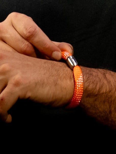Hand mit einem orangefarbenen, geflochtenen Armband und einer metallenen Verschlussklemme, die um das Handgelenk getragen wird.