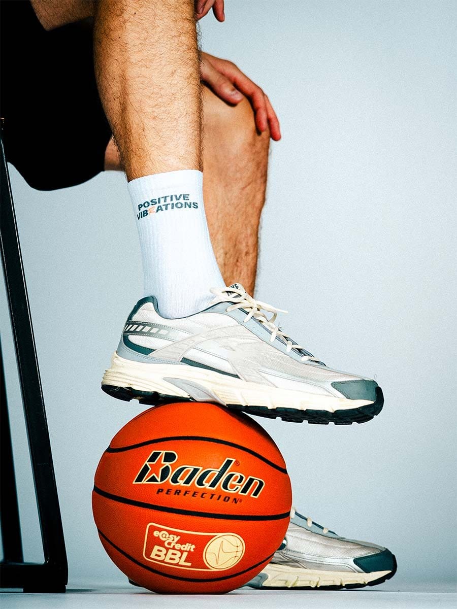 Ein Mann trägt weiße Sportschuhe, weiße Socken mit blauer Aufschrift, steht auf einem Basketball, der auf dem Boden liegt, und berührt sein Knie.