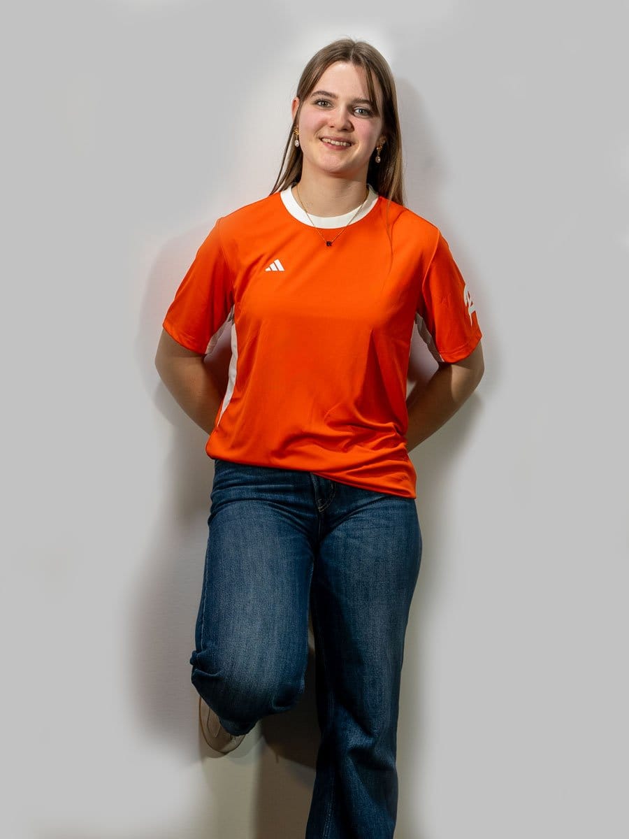 Junge Frau mit braunen Haaren, lächelnd, trägt orange Sportshirt mit weißen Details und blaue Jeans, steht vor grauer Wand, entspannt und freundlich.