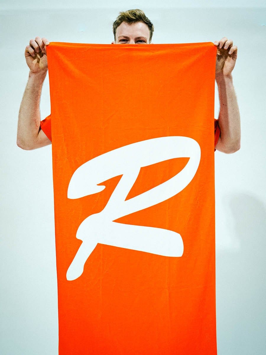 Ein Mann hält ein großes, orangefarbenes Tuch mit weißem Logo vor seinem Gesicht, sodass nur seine Augen und Hände sichtbar sind.