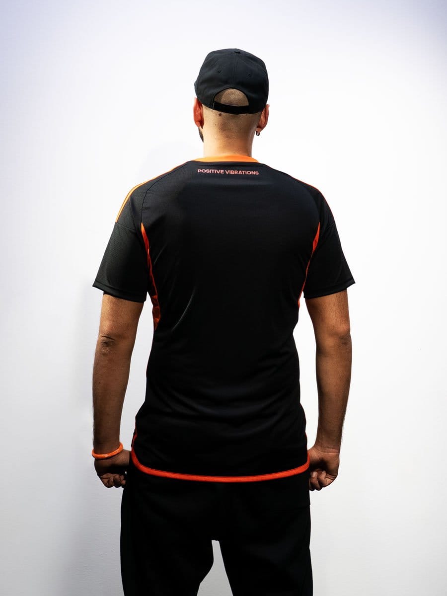 Mann trägt schwarze Sportbekleidung mit roten Akzenten, schwarze Mütze, steht vor weißer Wand, Rückenansicht, Text "Positive Vibrations" auf dem Rücken.