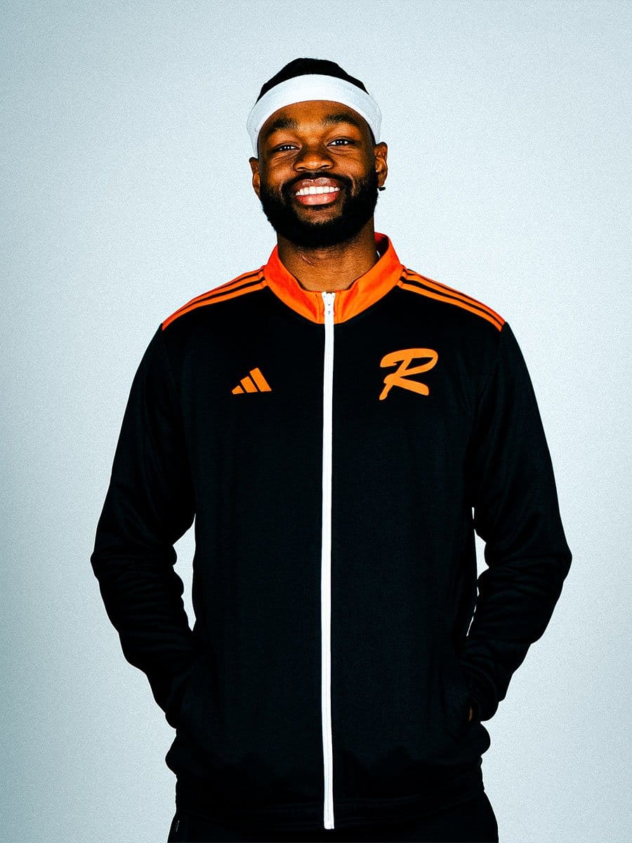 Lächelnder Mann in schwarzem Sportjacke mit orangefarbenen Streifen und Logo, trägt weißen Stirnband, steht vor hellem Hintergrund.