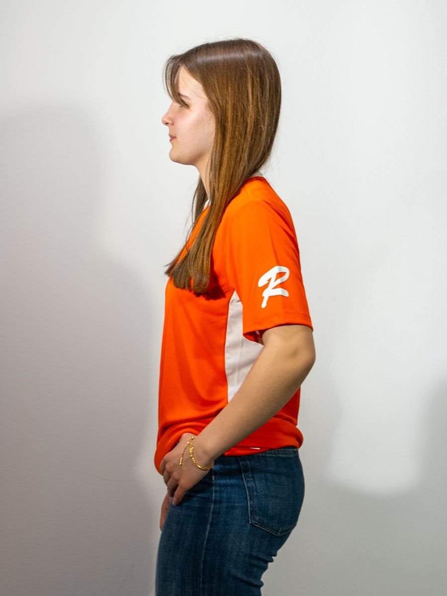 Seitliche Ansicht einer jungen Frau mit braunen Haaren, die ein rotes Sportshirt trägt und ihre rechte Hand in die Hosentasche steckt.