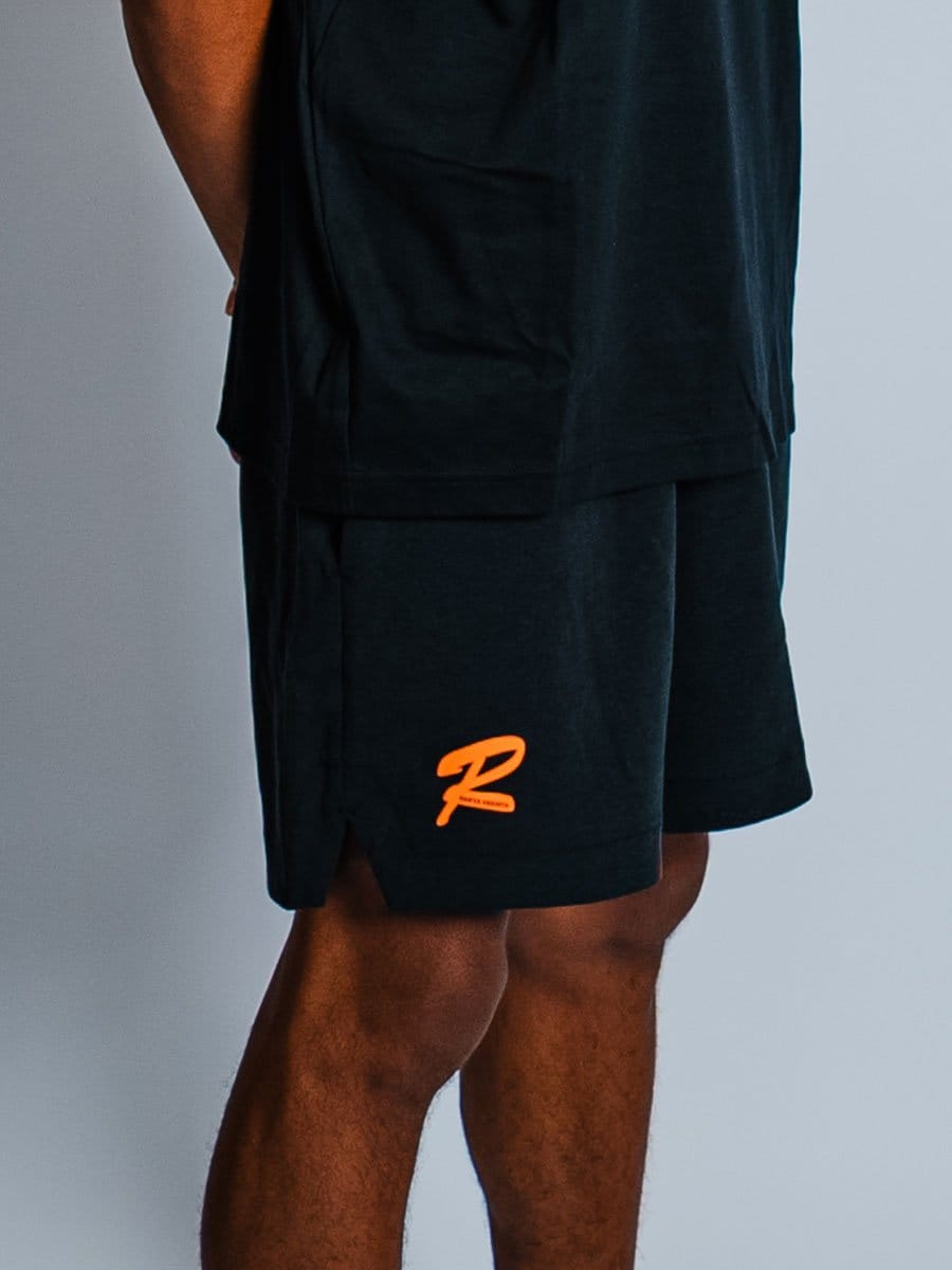 Schwarze Sportshorts mit orangefarbenem Logo auf der linken Seite, getragen von einer Person, die nur den unteren Rücken und die Beine zeigt.