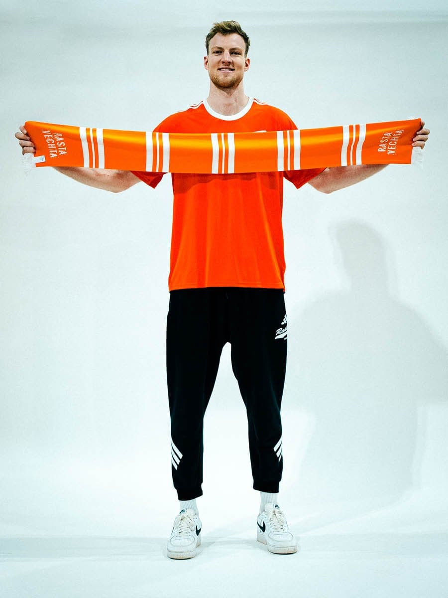 Junger Mann in rotem Sportshirt und schwarzen Adidas-Hosen hält einen orange-weißen Schal mit Text vor der Brust, steht vor weißem Hintergrund, lächelt.