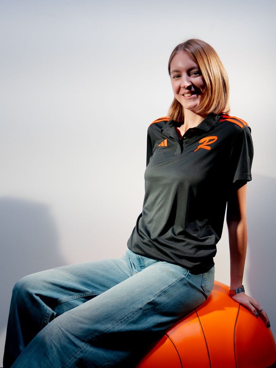Junge Frau sitzt auf einem großen, orangefarbenen Gymnastikball, trägt ein schwarzes Sportshirt mit orangefarbenen Akzenten und hat braunes, schulterlanges Haar.