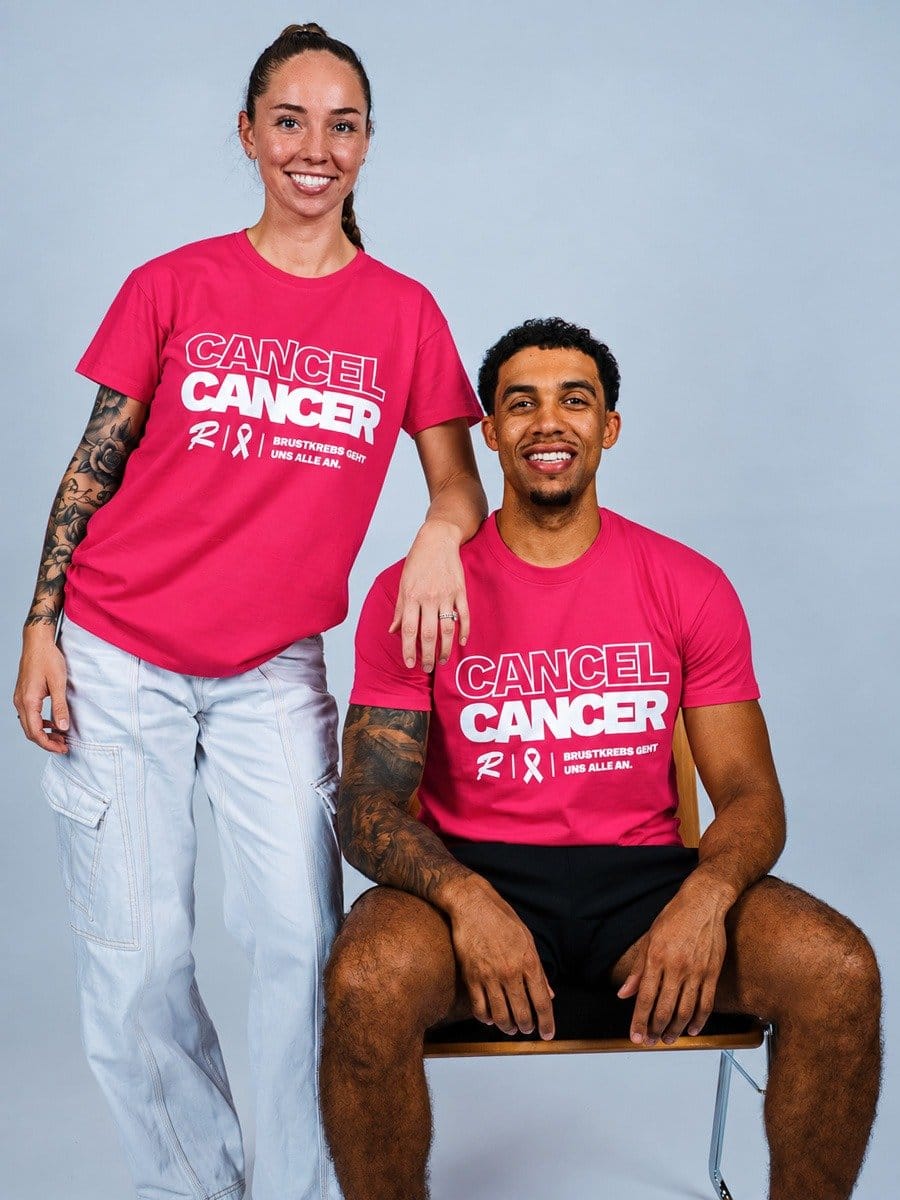 Zwei lächelnde Menschen, eine Frau und ein Mann, tragen pinke T-Shirts mit der Aufschrift "CANCEL CANCER" und einem Brustkrebs-Ribbon, vor hellem Hintergrund.