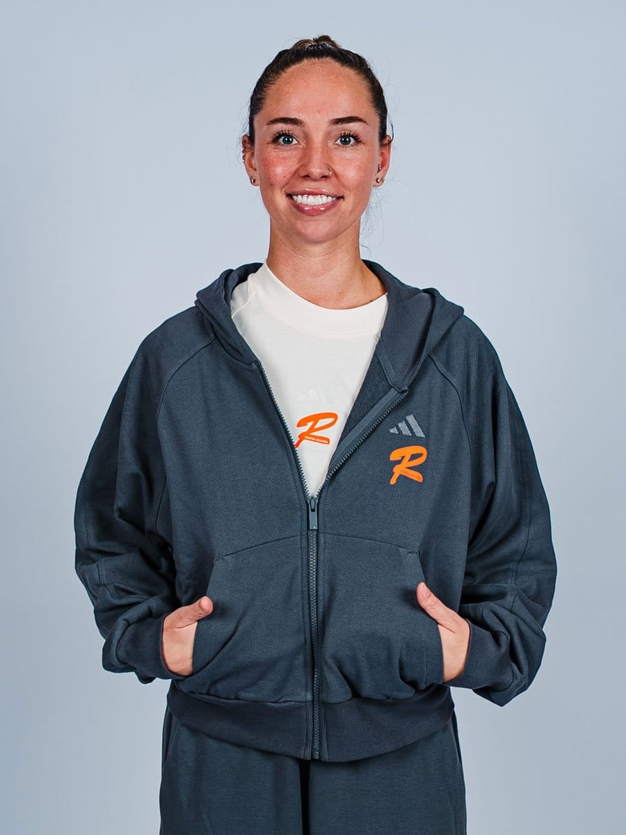 Junge Frau mit braunen Haaren in einem Zopf trägt eine dunkelblaue Kapuzenjacke mit orange und weißen Logos, lächelt und hat die Hände in den Taschen.