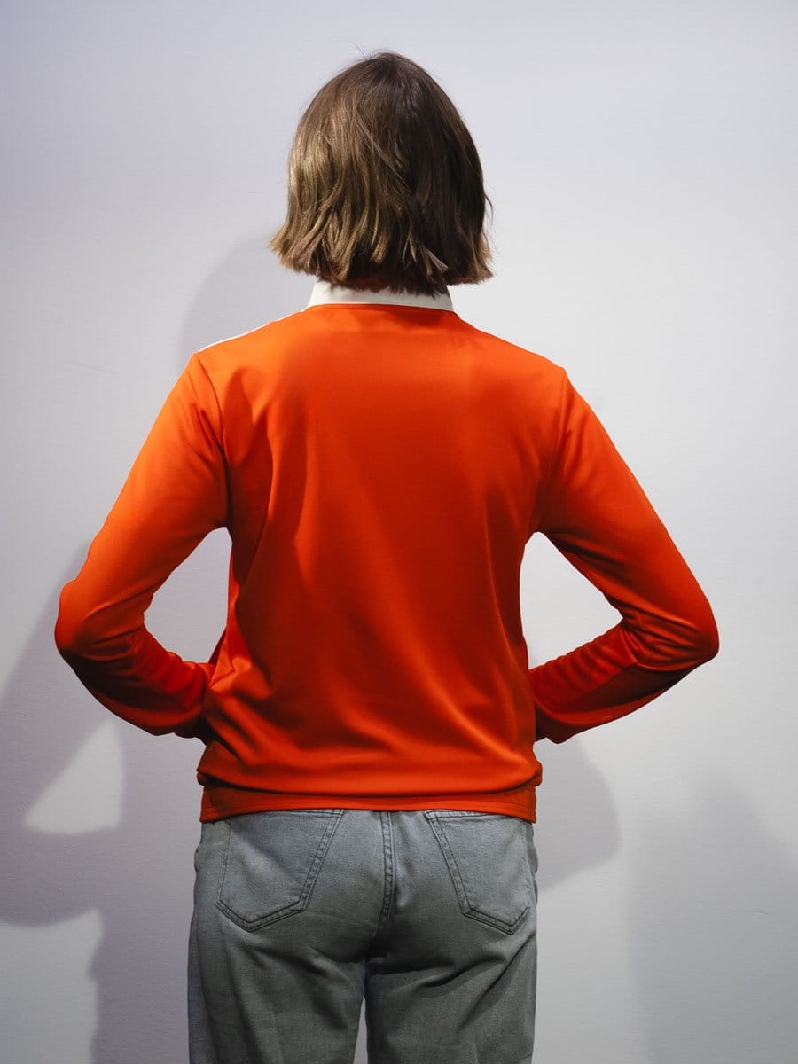Rückansicht einer Person mit braunen, schulterlangen Haaren, die ein rotes Oberteil und graue Jeans trägt, vor einer weißen Wand.