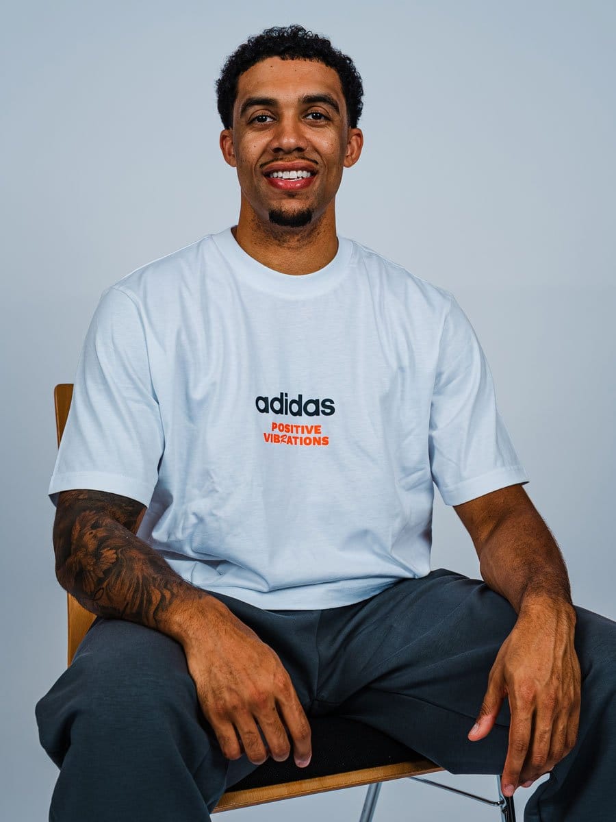 Ein lächelnder Mann mit dunklem, lockigem Haar sitzt auf einem Stuhl vor hellem Hintergrund, trägt ein weißes T-Shirt mit "adidas" und "Positive Vibrations" Schriftzug.