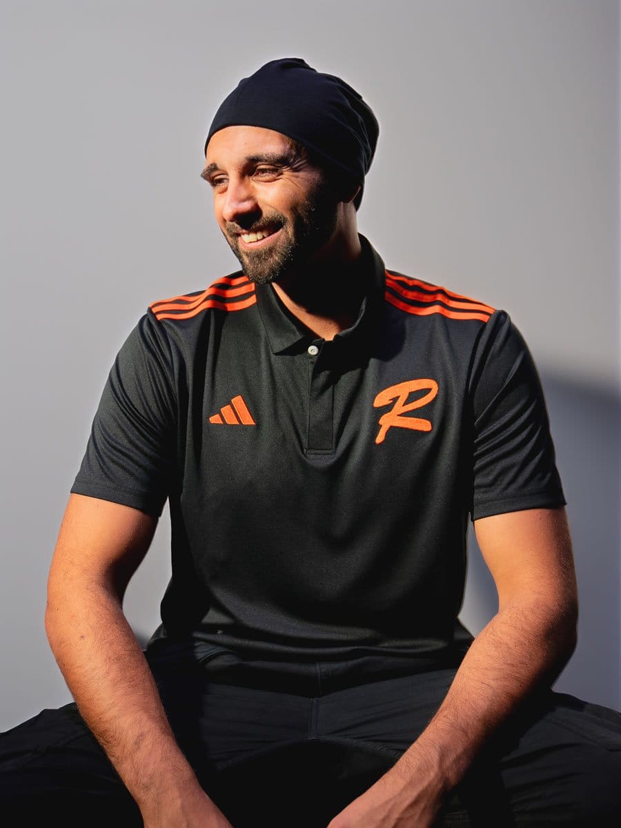 Mann mit dunklem Polo-Shirt mit orangefarbenen Streifen und Logo, schwarzer Beanie, lächelnd, sitzend vor hellem Hintergrund.