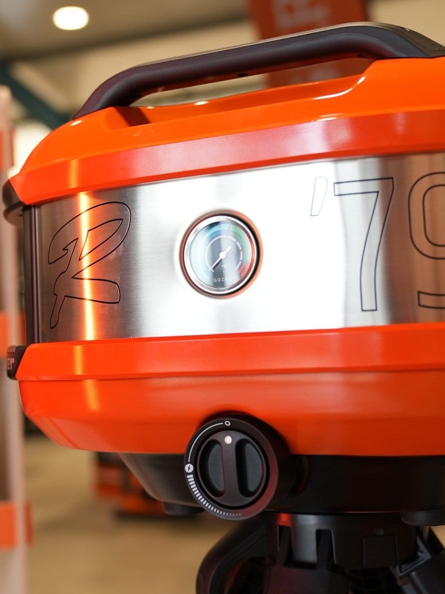 Runder, orange und silberner Grill mit Temperaturanzeige und Einstellknopf, auf einem schwarzen Standfuß.