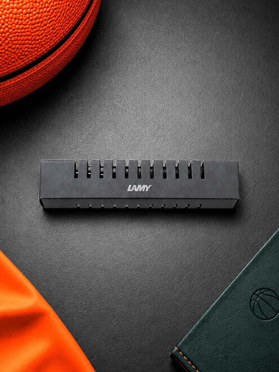 Schwarzer Basketball-Held mit der Aufschrift LAMY liegt auf einer schwarzen Oberfläche, umgeben von einem orangefarbenen Basketball, einem grünen Notizbuch und einem orangefarbenen Stoff.