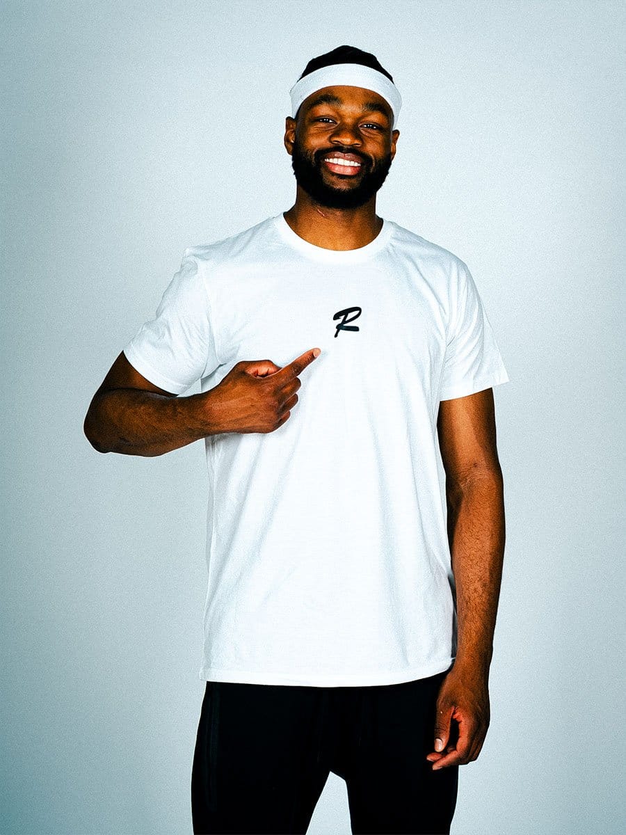 Ein lachender Mann mit dunklem Haar, Bart und dunkler Haut trägt ein weißes T-Shirt mit einem schwarzen "R" und zeigt auf das Logo.