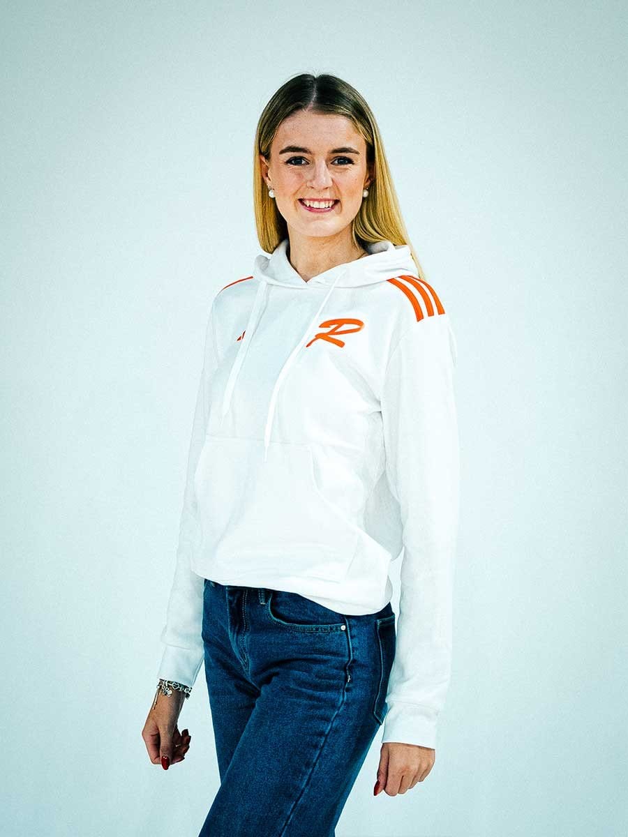 Junge Frau mit langem blondem Haar trägt weiße Kapuzenjacke mit roten Streifen und Logo, blaue Jeans, lächelt vor hellem Hintergrund.