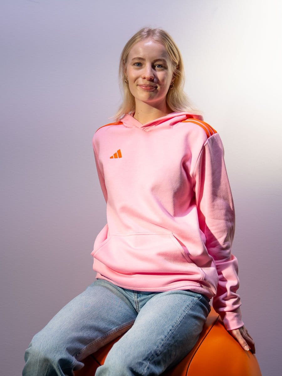 Junge Frau mit blonden Haaren sitzt auf einem orangefarbenen Ball, trägt eine pinke Hoodie mit orange Streifen an den Schultern, lächelt und blickt in die Kamera.