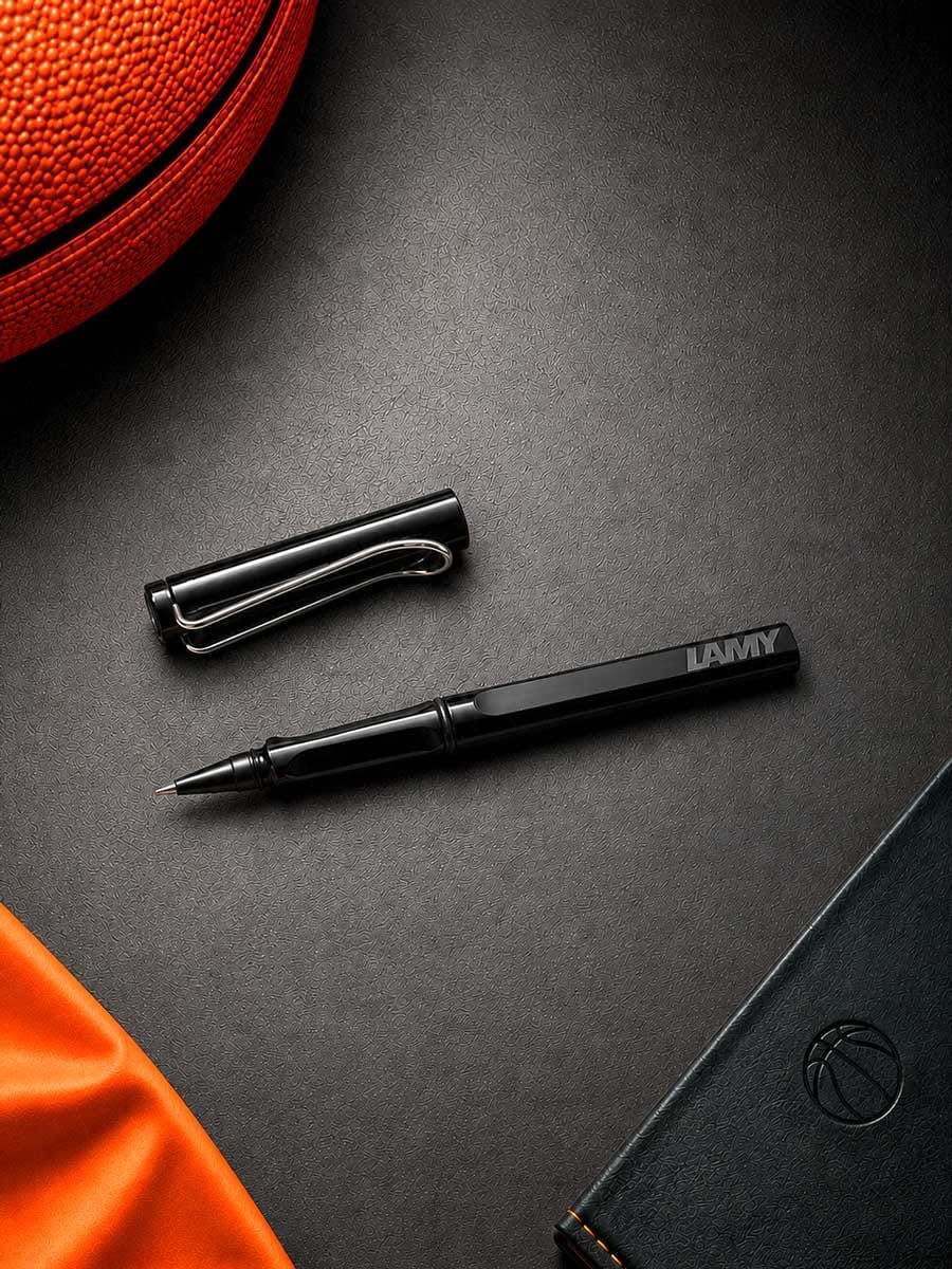 Schwarzer LAMY Füllfederhalter mit abnehmbigem Clip liegt auf schwarzer Oberfläche neben einem schwarzen Notizbuch und einem roten Basketball.