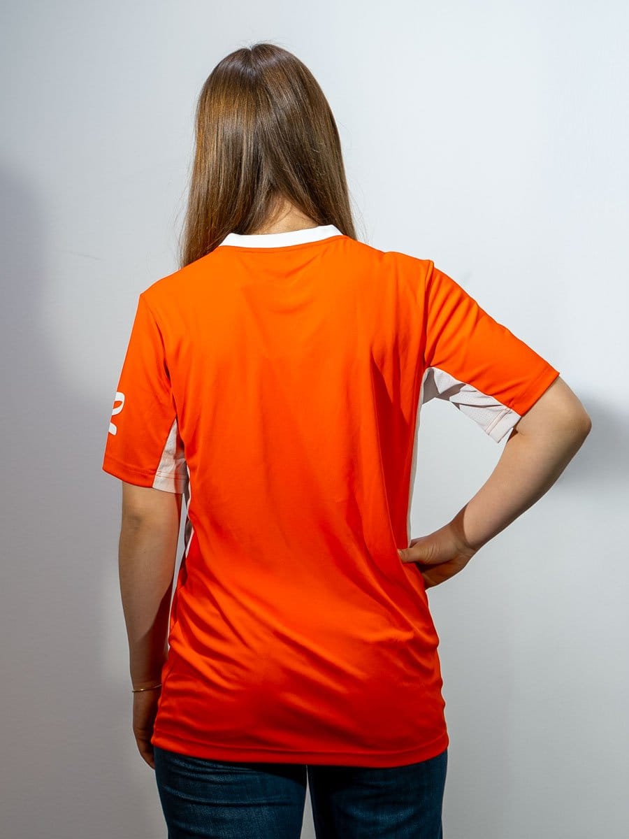 Rückansicht einer Frau mit braunen Haaren, die ein orangefarbenes Sportshirt trägt, das an den Seiten weiß abgesetzt ist, und ihre rechte Hand auf die Hüfte stützt.