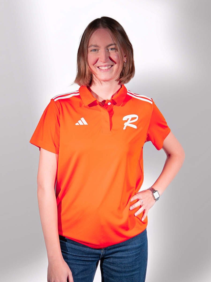 Junge Frau mit braunen Haaren, lächelnd, trägt eine orange Sportjacke mit weißen Streifen und einem weißen "R" auf der Brust, steht vor hellem Hintergrund.