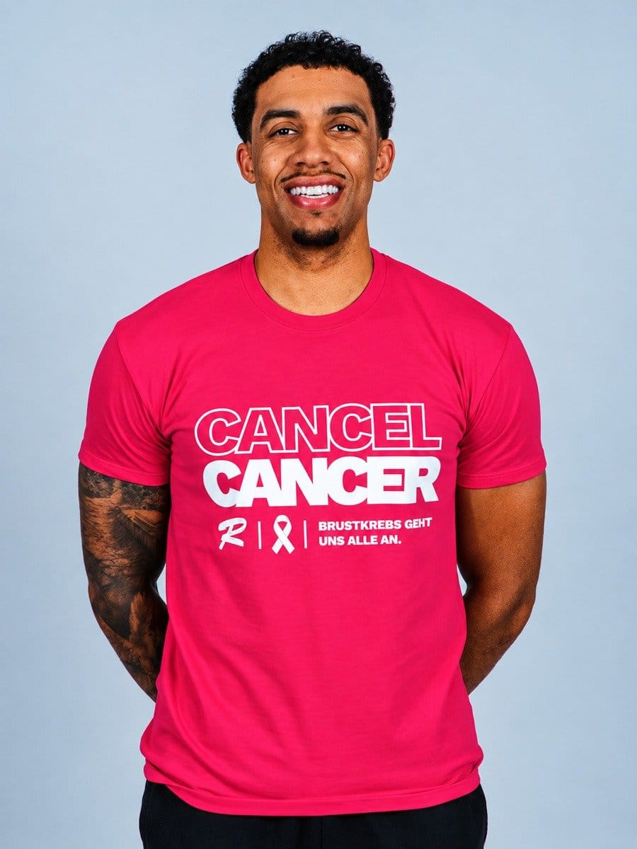 Mann trägt pinkes T-Shirt mit weißem Text: "CANCEL CANCER RIX Brustkrebs geht uns alle an" vor hellem Hintergrund.