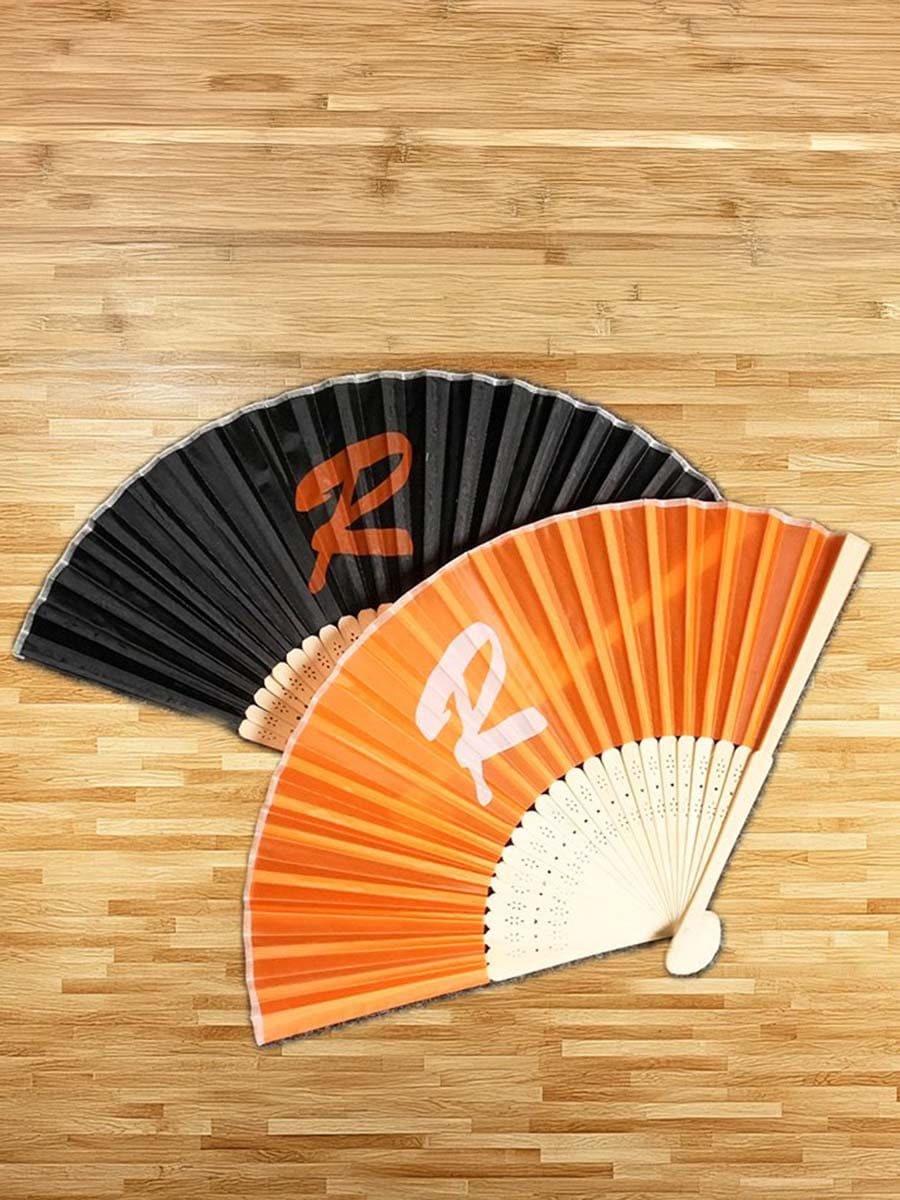 Zwei faltbare Handfächer, einer schwarz mit orangeem "R"-Logo, der andere orange mit weißen "R"-Logo, liegen auf einem Holztisch.
