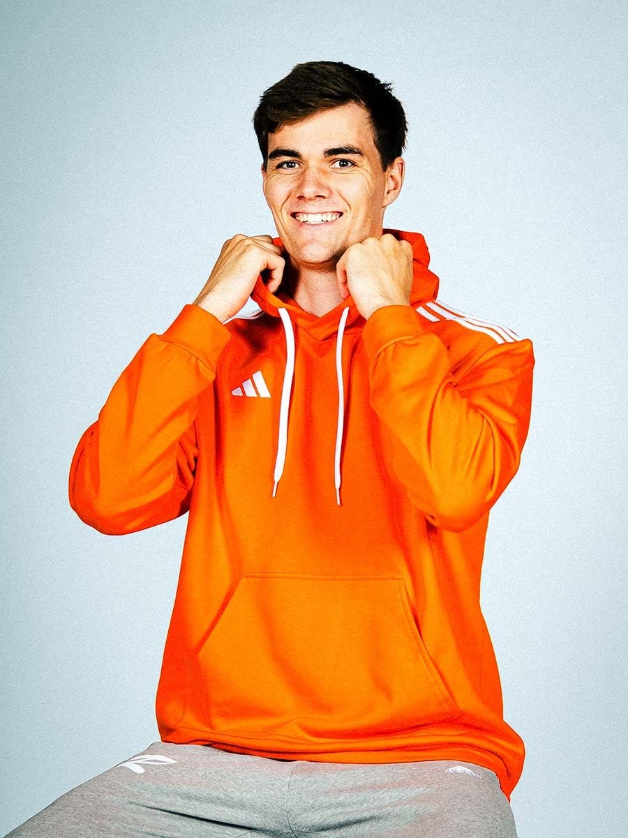 Junger Mann in orange Adidas-Sportjacke mit weißen Streifen, lächelnd, hält die Kapuze mit beiden Händen, sitzt vor hellem Hintergrund.