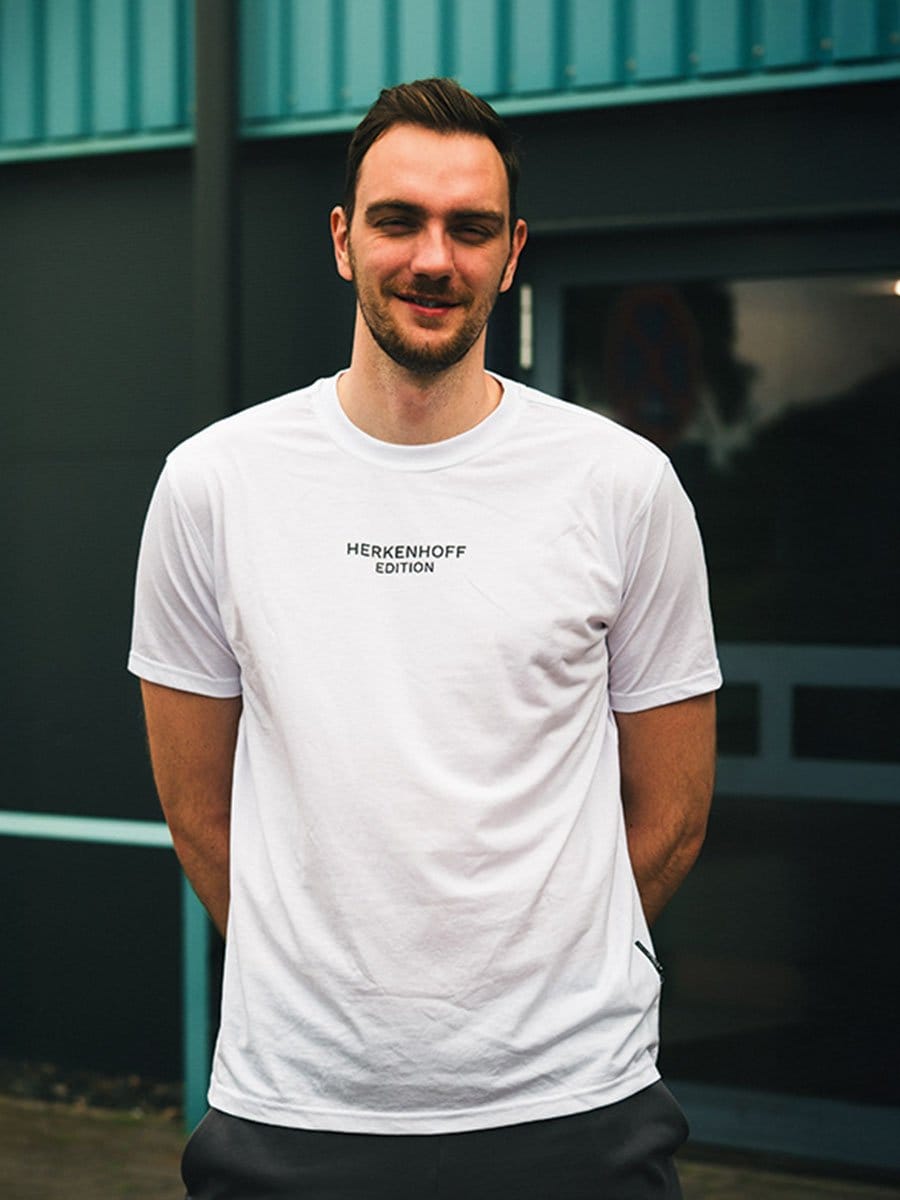 Junger Mann mit dunklem Haar und Bart, trägt ein weißes T-Shirt mit "HERKENHOFF EDITION" auf der Brust, steht vor einem modernen Gebäude.