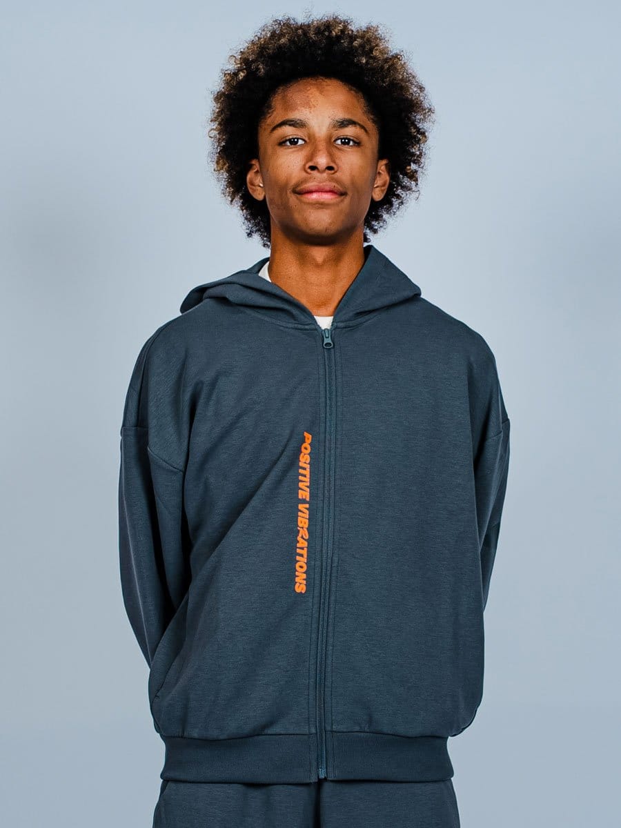 Junge Person trägt eine dunkelblaue Kapuzenjacke mit orange Schriftzug "POSITIVE VIBRATIONS" auf der linken Seite, steht vor hellem Hintergrund.