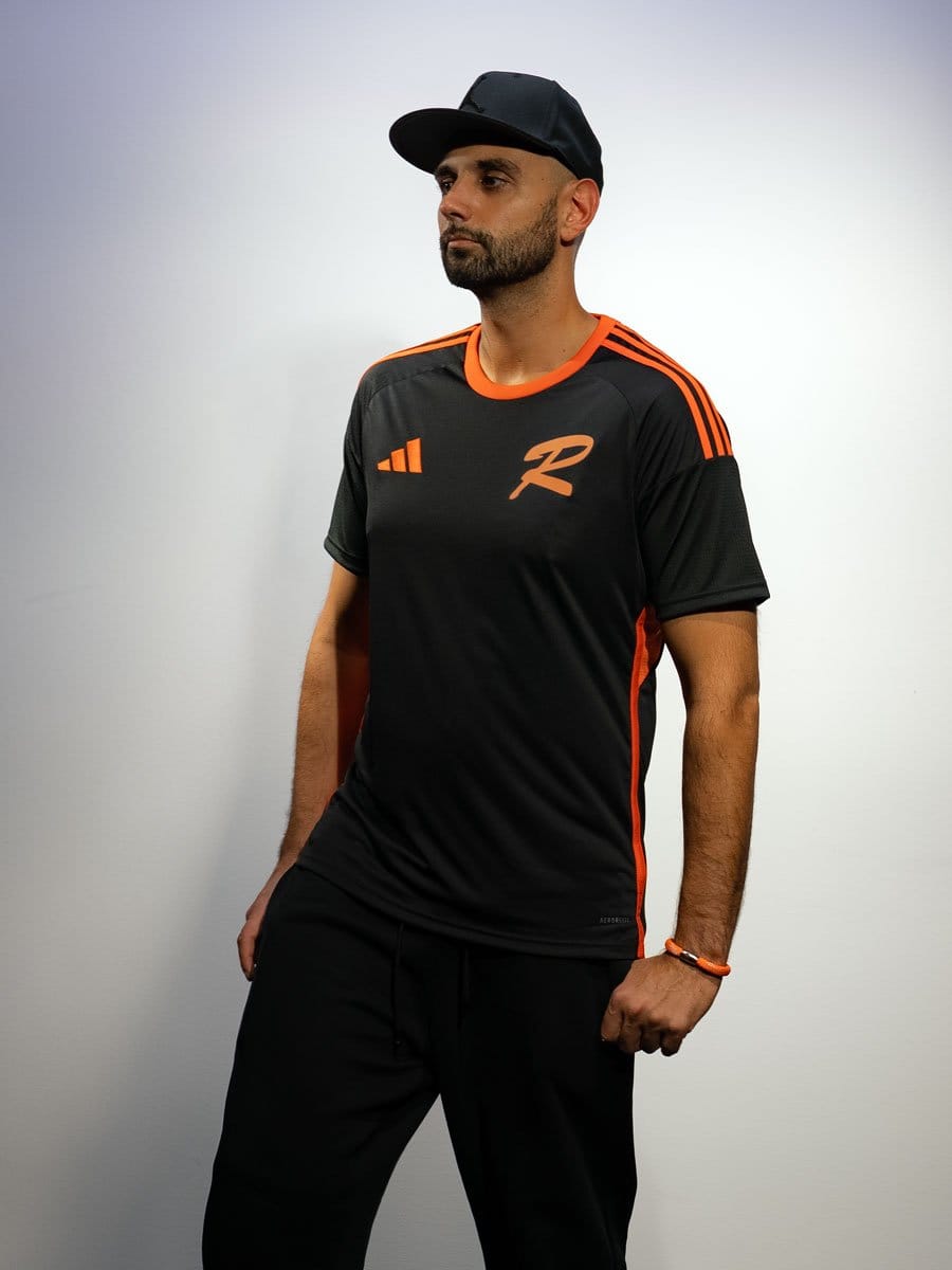 Mann in schwarzem Sportshirt mit orangefarbenen Details, schwarzer Cap, schwarze Hose, steht vor weißer Wand, Blick nach links, entspannt.