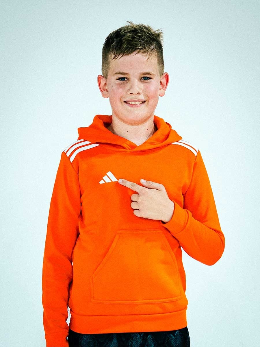Junge trägt orange Adidas-Hoodie, zeigt auf das Logo auf der Brust, lächelt vor weißem Hintergrund.