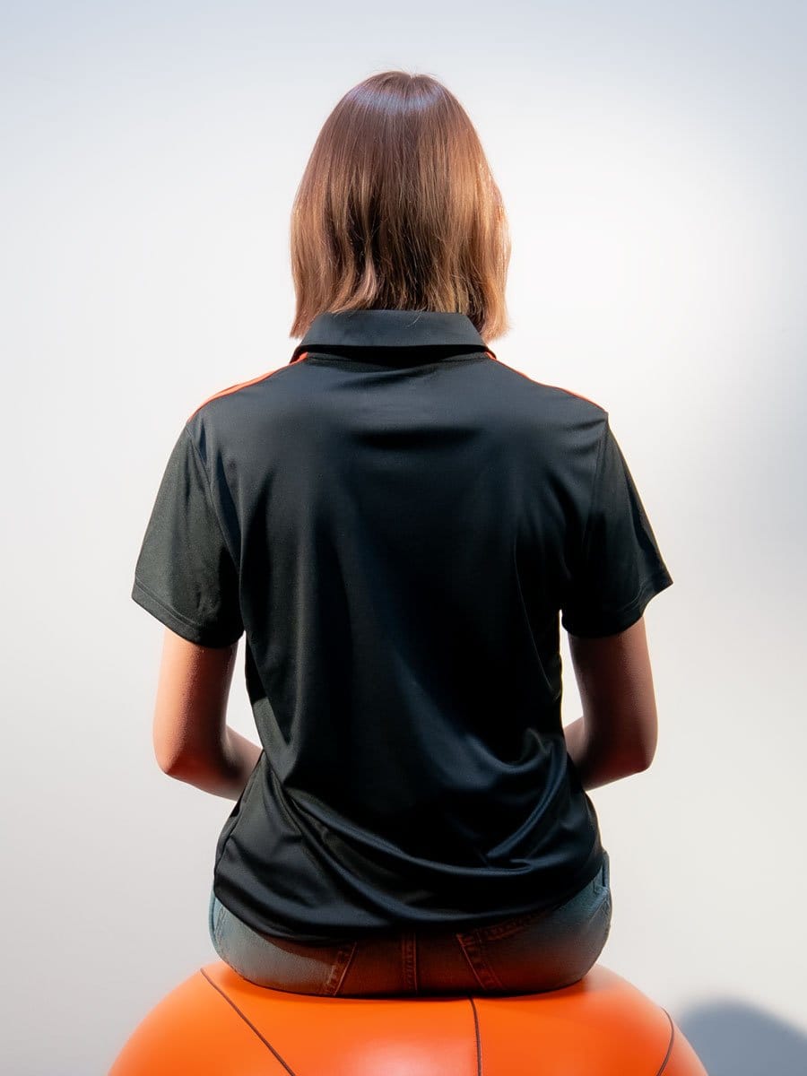 Rückansicht einer Frau mit braunen, schulterlangen Haaren, sitzend auf einem orangefarbenen Gymnastikball, trägt ein schwarzes Sportshirt mit roten Akzenten.