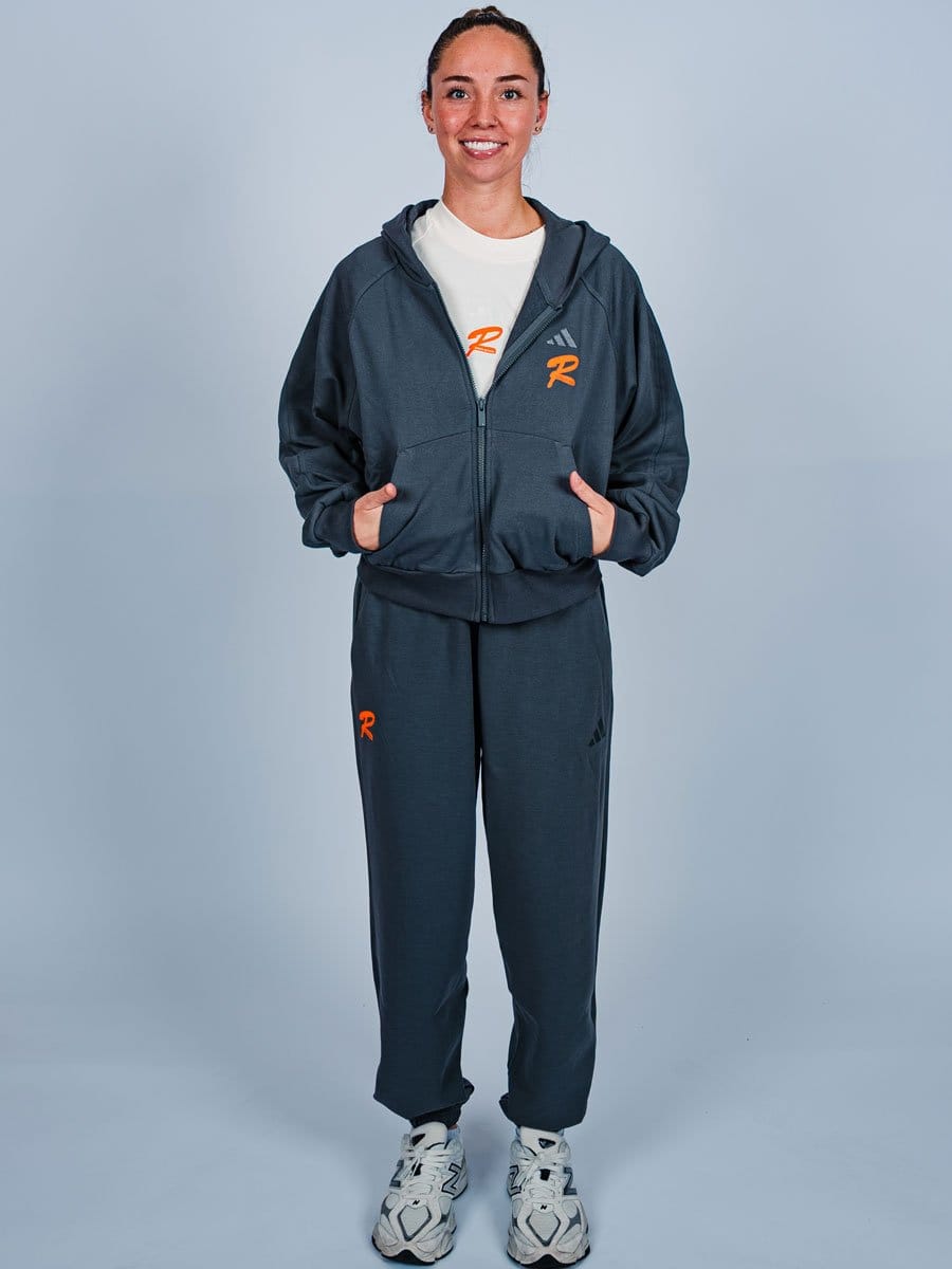 Junge Frau in dunkler Sportbekleidung mit weißen Turnschuhen, lächelnd, steht vor hellem Hintergrund, trägt eine Kapuzenjacke und Jogginghose.
