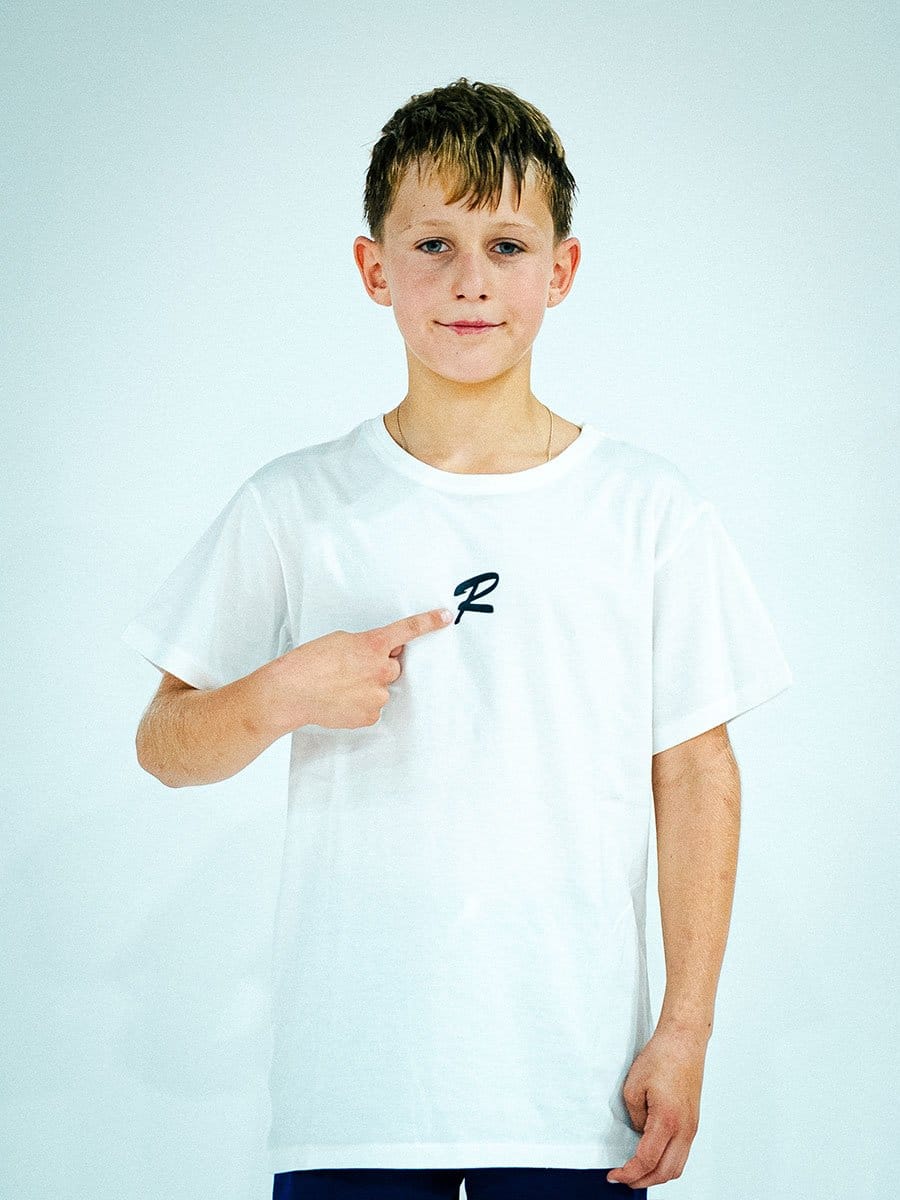 Junge trägt weißes T-Shirt mit schwarzem Logo, zeigt mit Finger auf das Logo, steht vor hellem Hintergrund, lächelt leicht.