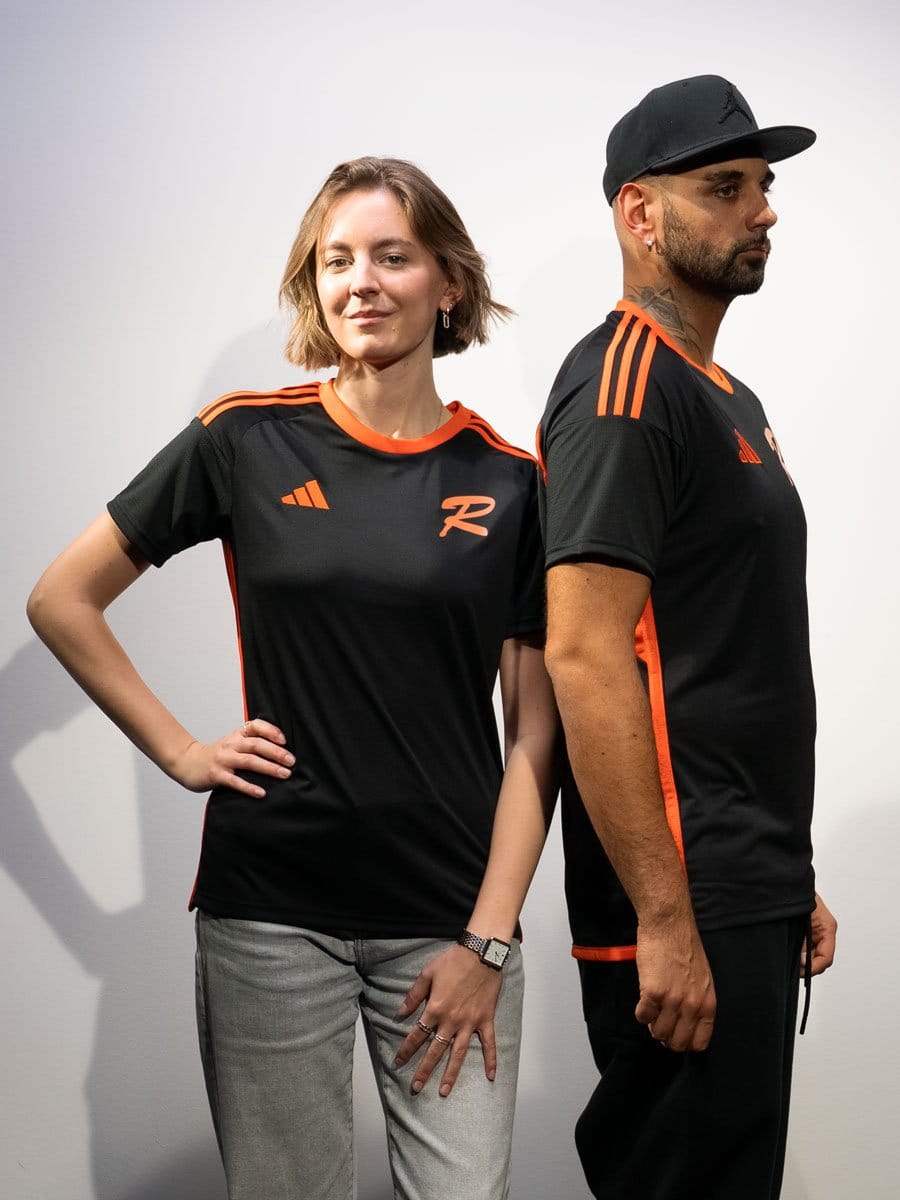 Zwei Menschen in schwarzen Sport-T-Shirts mit orangefarbenen Akzenten, eines mit kurzen braunen Haaren und das andere mit schwarzen Haaren und Cap, stehen nebeneinander.