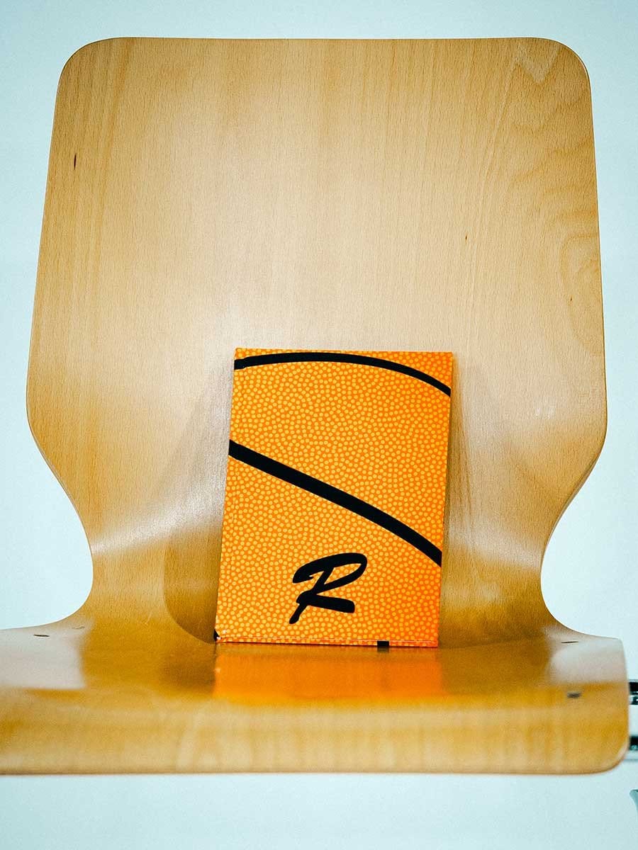 Ein orangefarbenes, quadratisches Produkt mit schwarzen Linien und einem schwarzen "R" auf einem Holzstuhl.