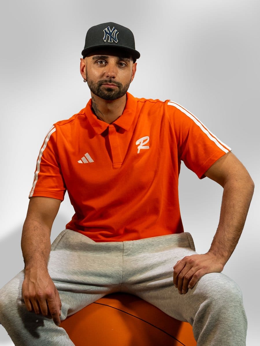 Mann sitzt auf einem orangefarbenen Ball, trägt ein orangefarbenes Polo-Shirt mit weißen Streifen an den Schultern und eine schwarze Baseballcap mit NY-Logo.