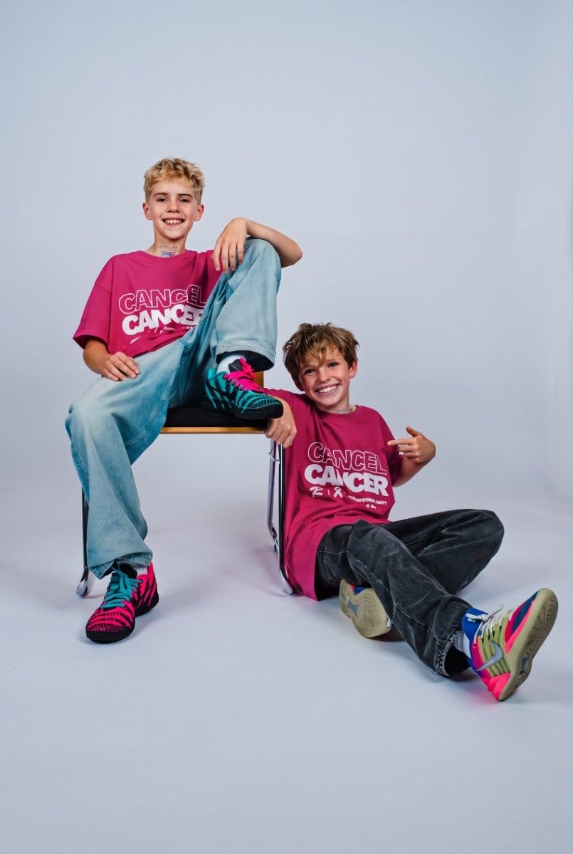 Zwei lachende Kinder in pinken T-Shirts mit "Cancel Cancer"-Aufdruck, eines sitzt auf einem Stuhl, das andere liegt auf dem Boden, beide tragen bunte Turnschuhe.