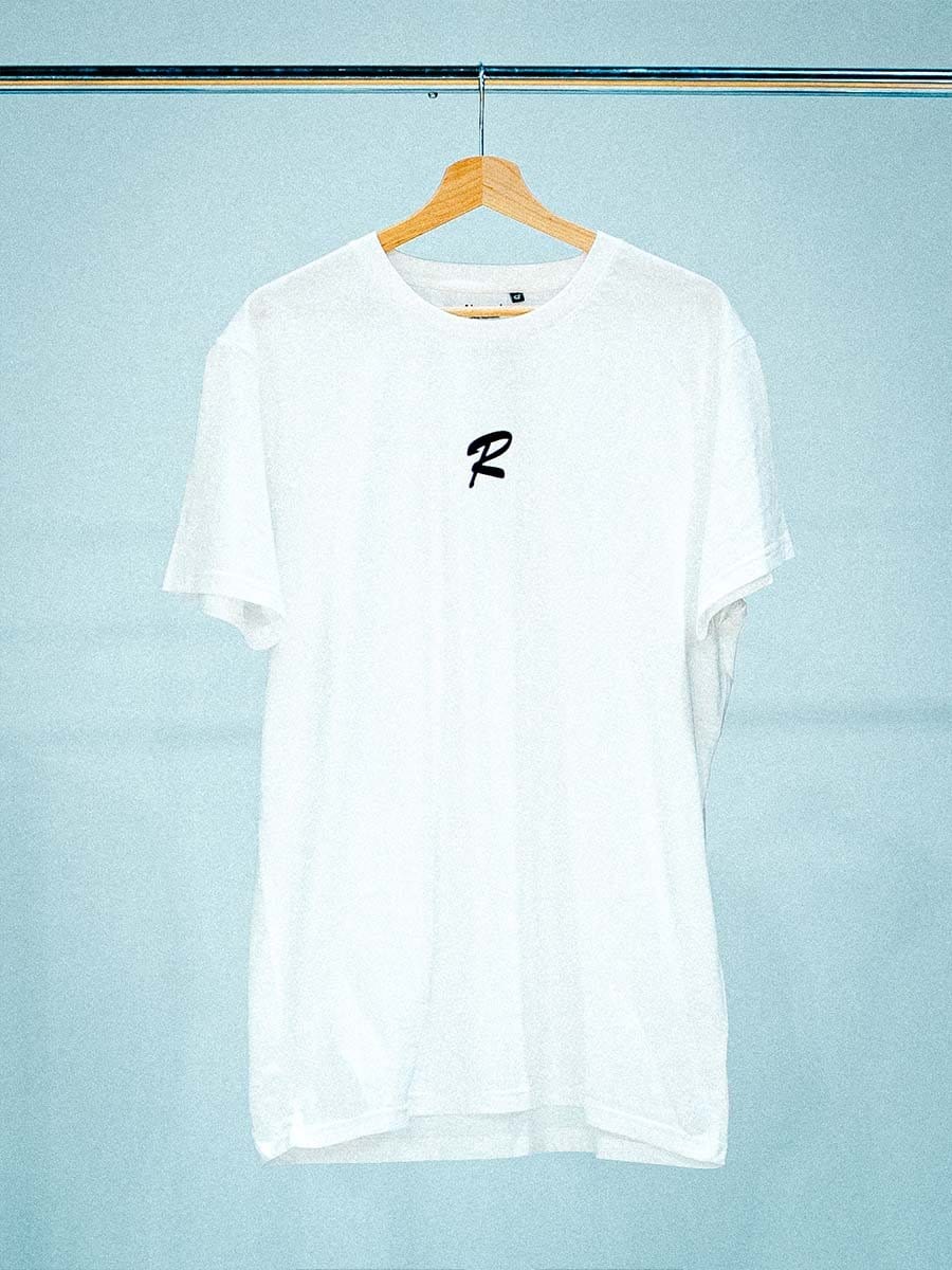 Weißes T-Shirt mit schwarzem Buchstaben "R" in der Mitte, hängt an einem Holzkleiderbügel an einer Stange vor einem hellblauen Hintergrund.
