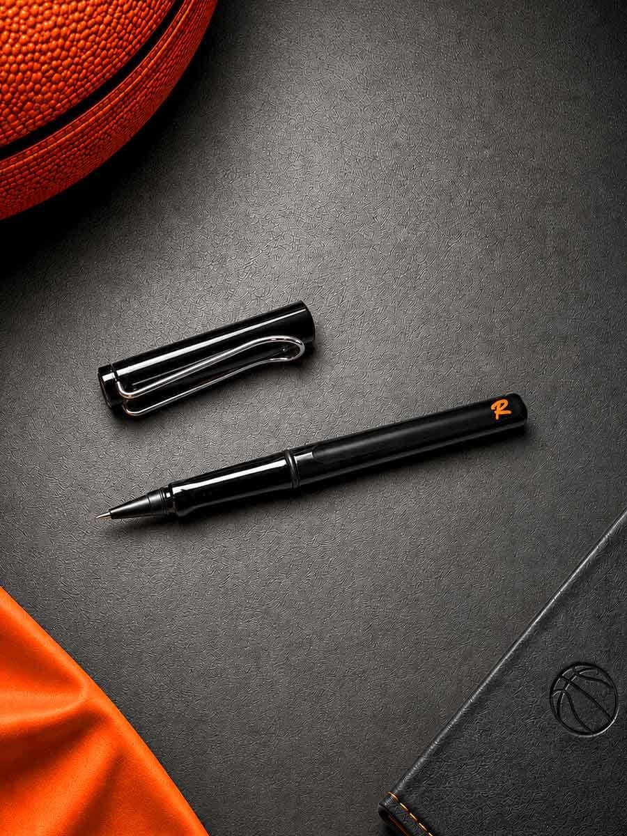 Schwarzer Kugelschreiber mit orangeem Logo, offen mit herausgezogenem Schreibkopf, auf schwarzer Oberfläche mit rotem Stoff im oberen linken Bereich.
