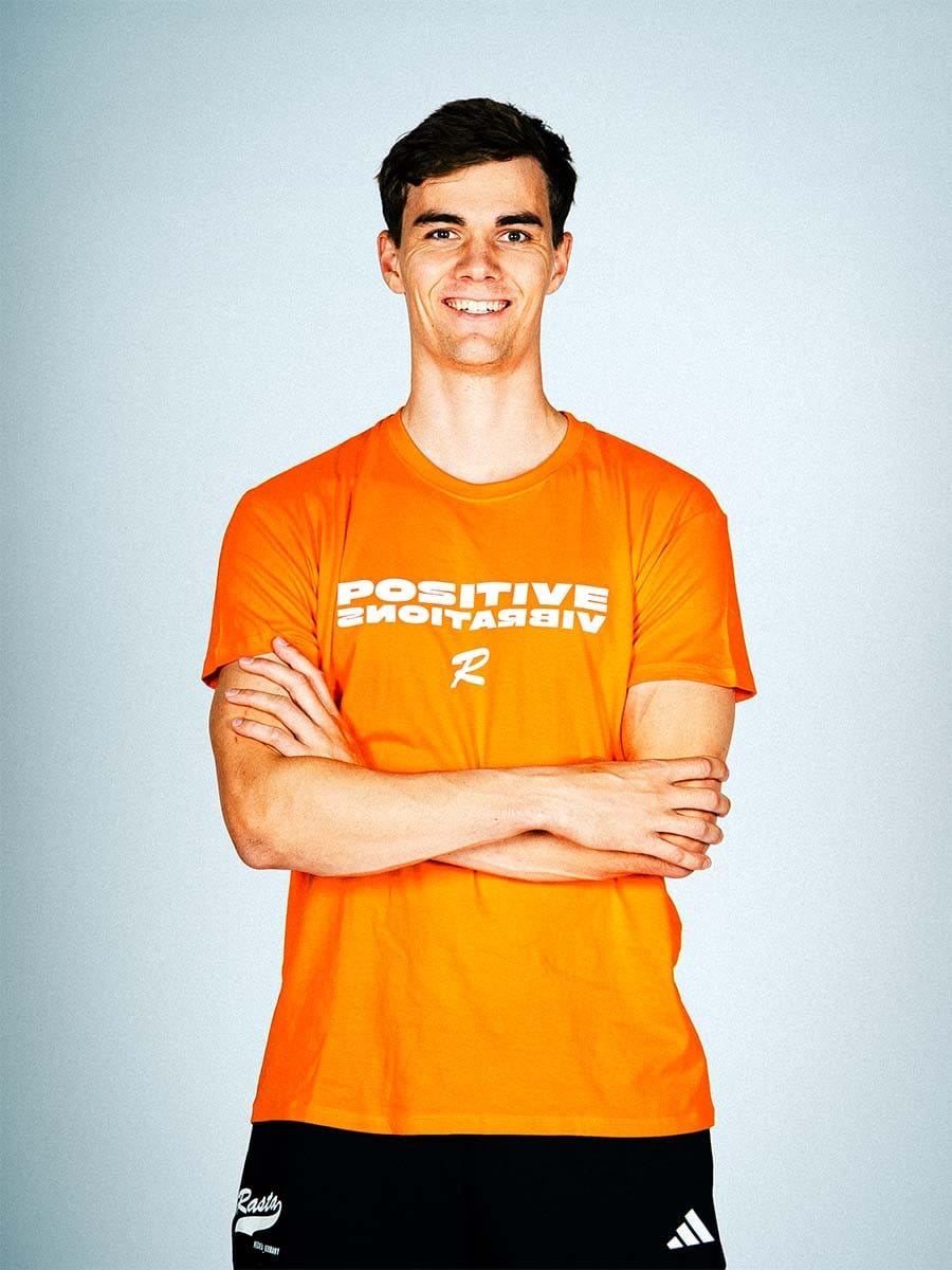Junge Mann trägt orange T-Shirt mit weißen Text "POSITIVE VIBRATIONS" und schwarzen Adidas-Hosen, steht mit verschränkten Armen vor hellem Hintergrund.