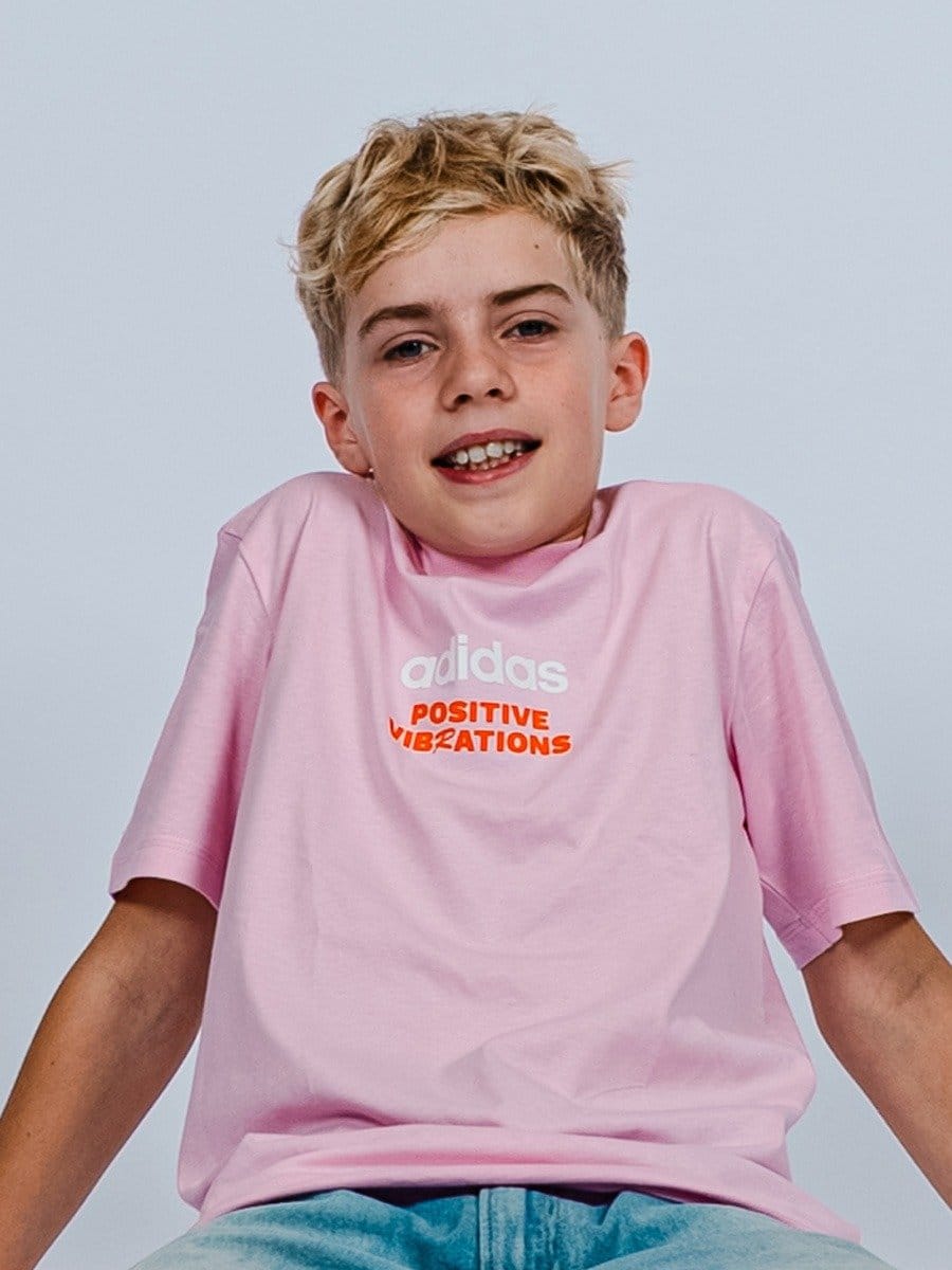 Junge trägt ein pinkes T-Shirt mit weißen und roten Aufdrucken, sitzt entspannt, lächelt leicht und blickt in die Kamera. Hintergrund hell und schlicht.