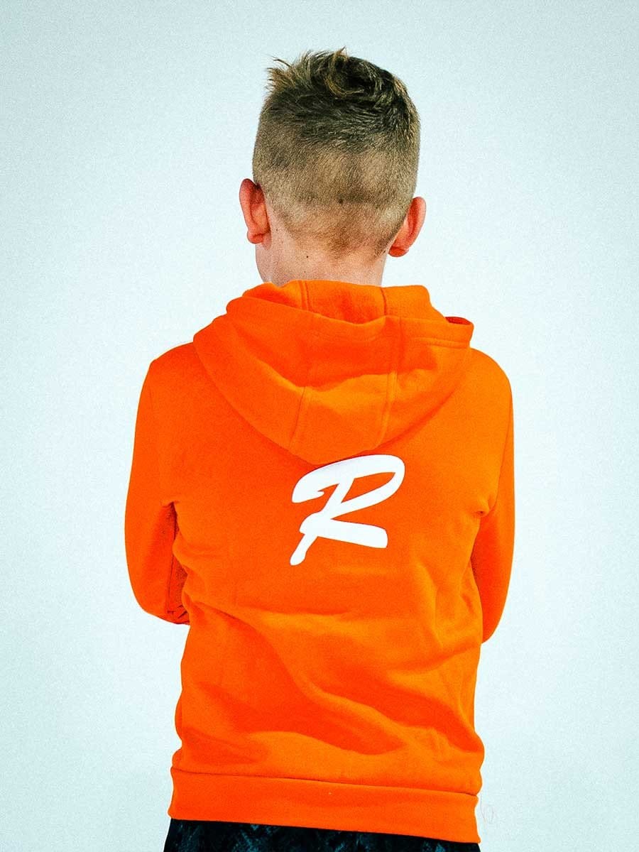 Rückenansicht eines Jungen in einer orange Hoodie mit weißem Logo auf dem Rücken, kurz geschnittene Haare, vor hellem Hintergrund.