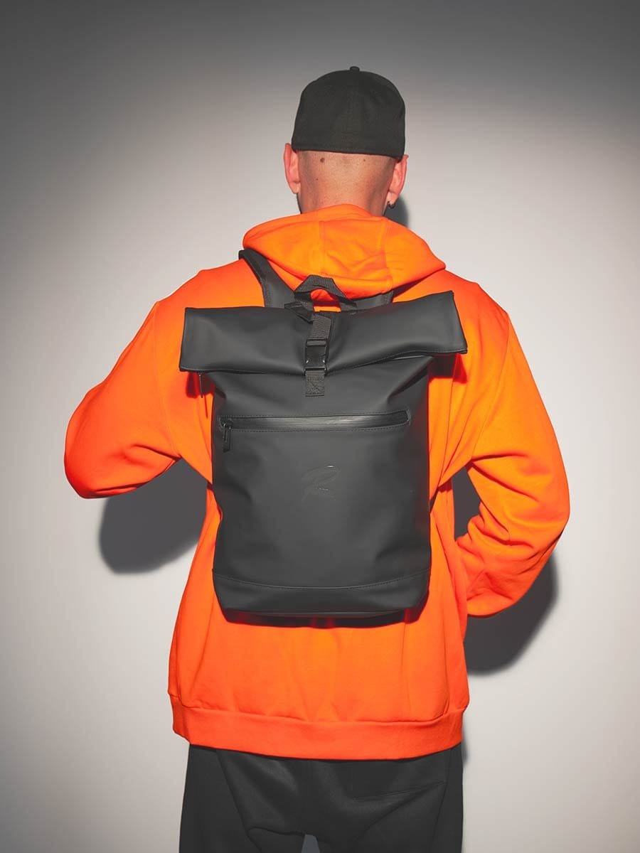 Mann mit schwarzem Cap, orangeem Hoodie und schwarzem Rucksack, der von hinten fotografiert wird, vor einer grauen Wand.