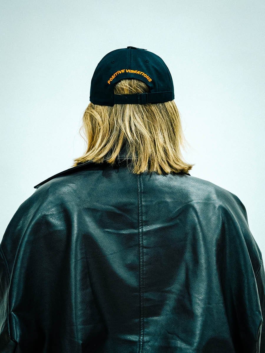 Person mit Schulterlänge blondem Haar, schwarze Lederjacke und schwarzer Baseballcap mit orangeener Aufschrift "POSITIVE VIBRATIONS" von hinten gesehen.