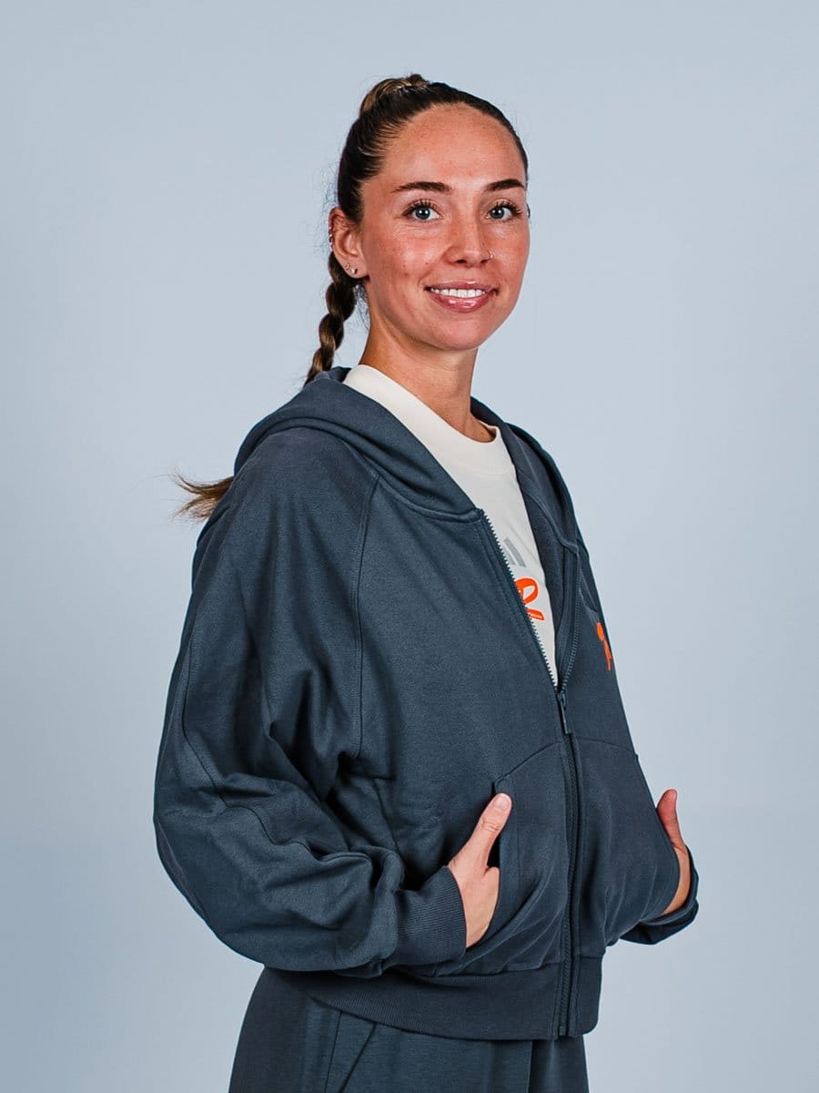 Junge Frau mit braunem Zopf, lächelnd, trägt eine dunkelblaue Kapuzenjacke und ein weißes T-Shirt, steht vor hellem Hintergrund.
