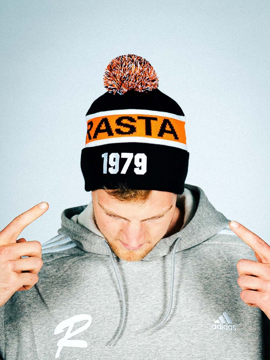 Junger Mann trägt grauen Hoodie und schwarze Mütze mit orange-weißer Banderole, der Aufschrift "RASTA" und "1979" in Weiß, zeigt auf die Mütze.