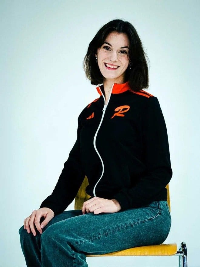 Junge Frau mit dunklen Haaren sitzt auf einem Stuhl, trägt eine schwarze Jacke mit orangefarbenen Details und lächelt.